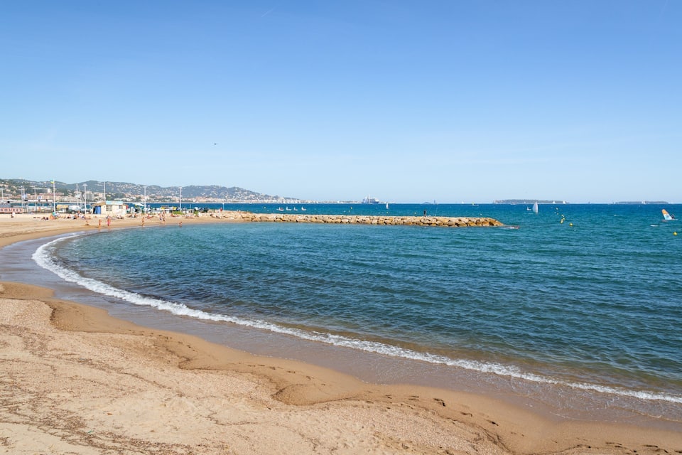 A sandy beach with clear blue water, a rocky breakwater, and distant buildings.