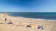A sandy beach with people sunbathing and swimming, a rocky breakwater, and sailboats in the distance.