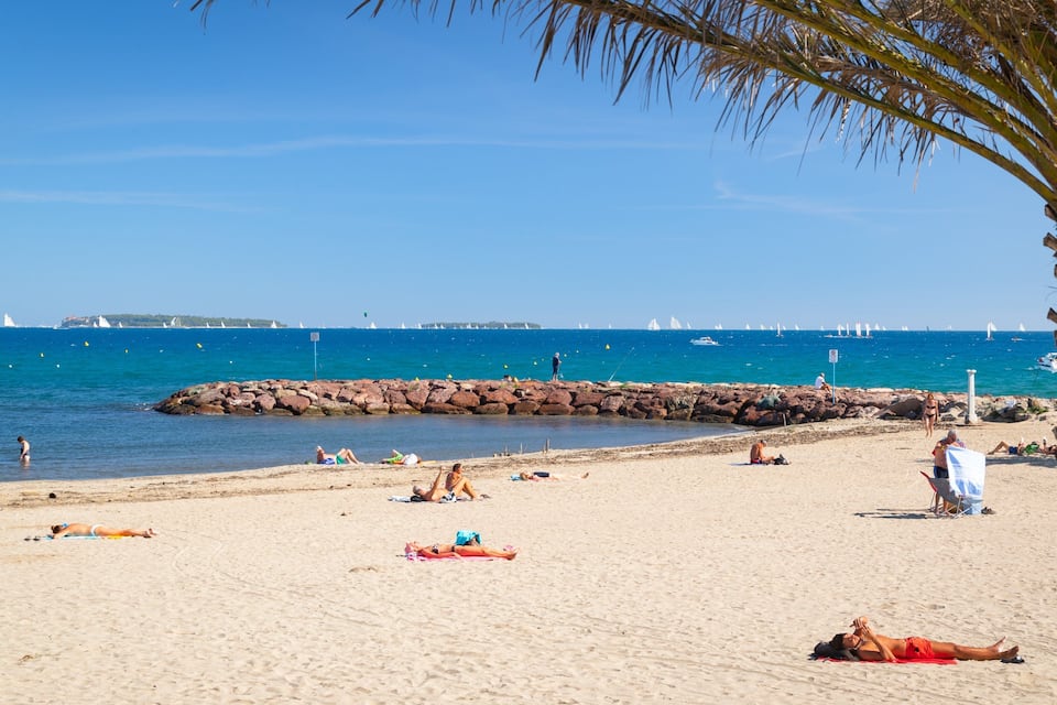 Ein sandiger Strand mit badenden und sonnenden Menschen, ein Felssteg und Segelboote in der Ferne.