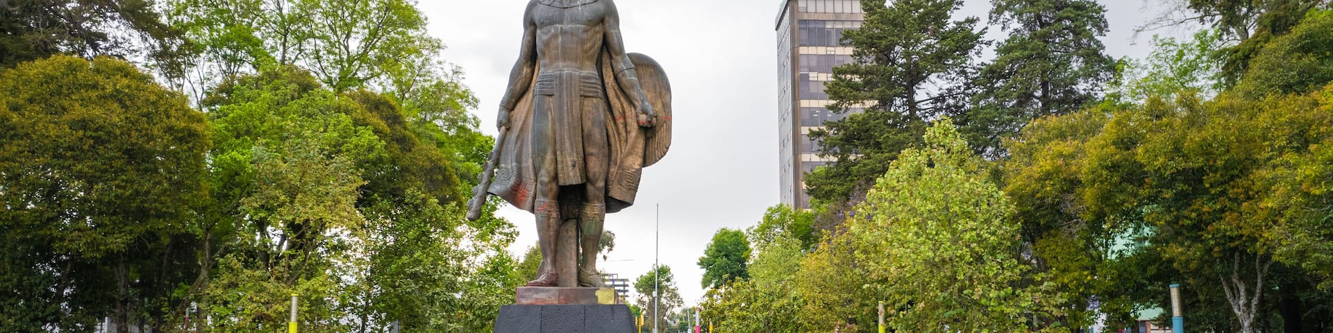 A statue of a woman holding a shield in a park with trees and a building in the background.
