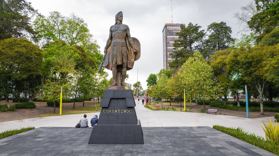 A statue of a woman holding a shield in a park with trees and a building in the background.