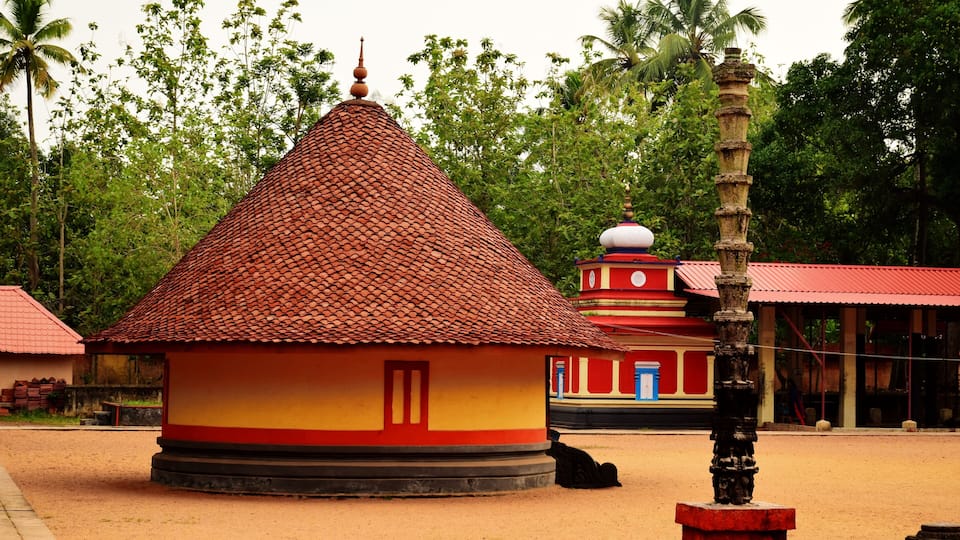 A traditional building with a red-tiled roof and a small door, surrounded by greenery and palm trees.