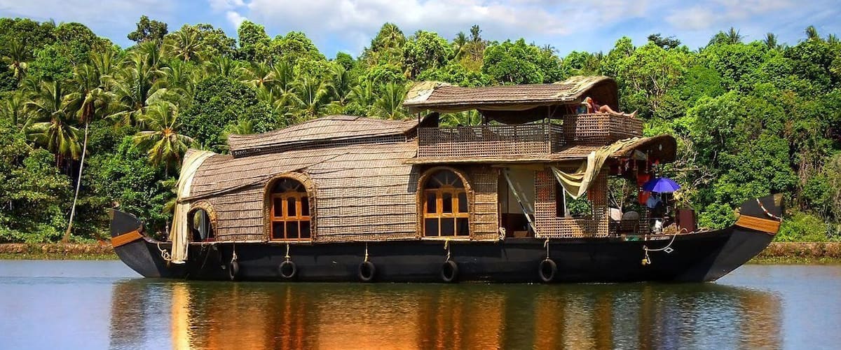 A traditional houseboat on water with a thatched roof, surrounded by lush greenery and palm trees.