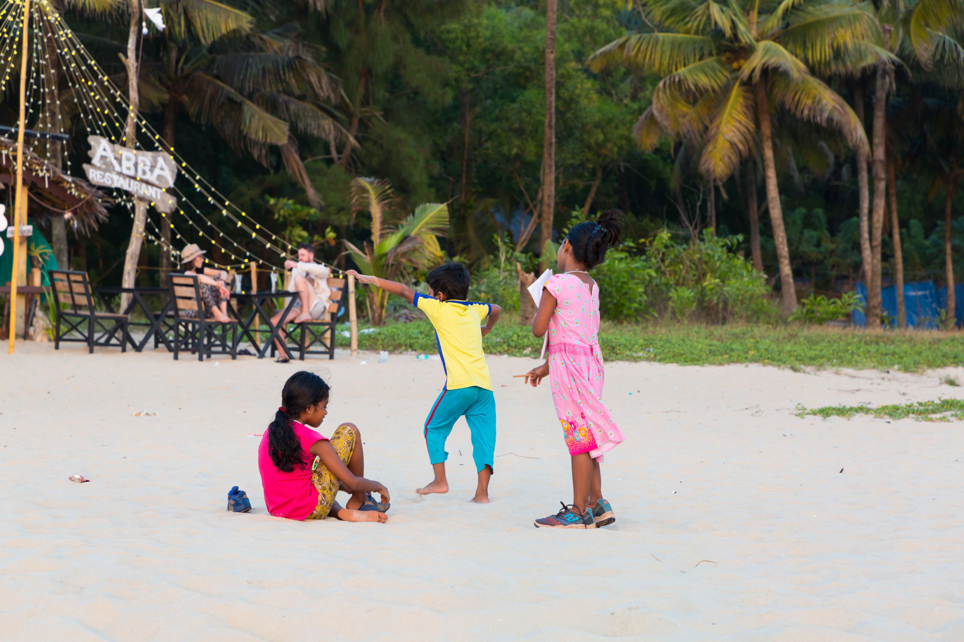 Children playing on a sandy beach with palm trees and a restaurant sign in the background.