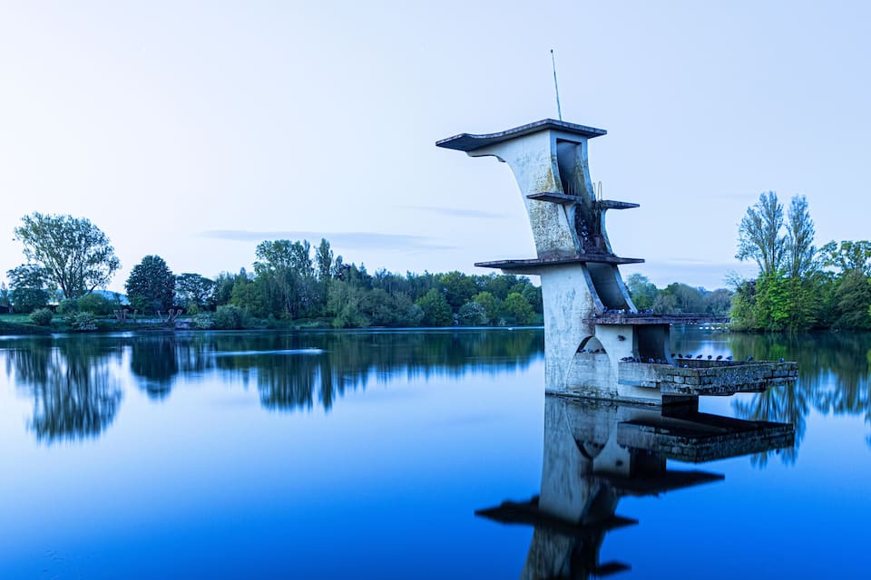 A unique tower structure with a reflection in a calm lake, surrounded by trees and greenery.