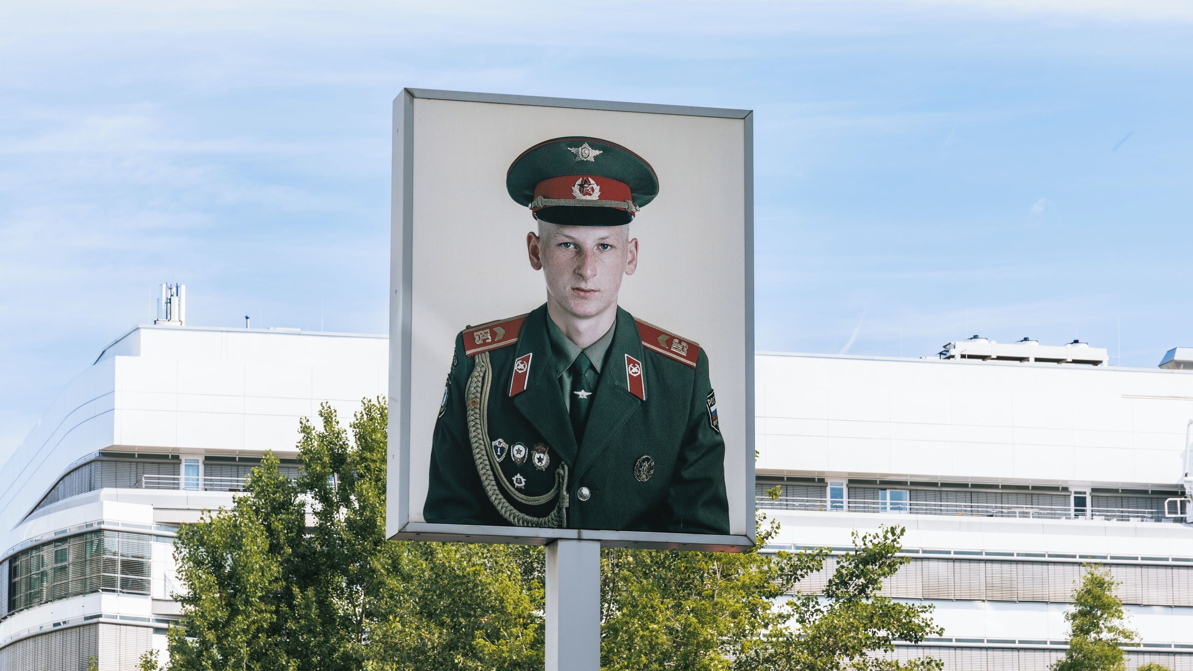 A portrait of a person in a military uniform with medals, standing in front of a modern building with a clear sky.