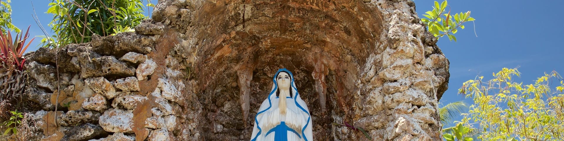 Una estatua de una mujer con una túnica azul y blanca, ubicada en un hueco rocoso rodeado de follaje.