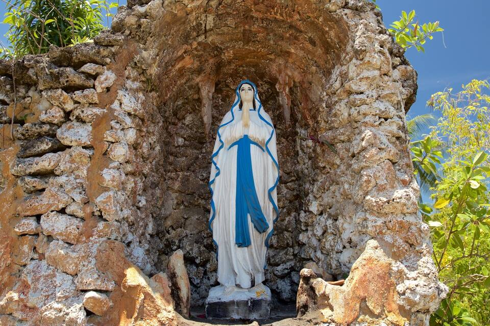 A statue of a woman in a blue and white robe, standing in a rocky niche surrounded by greenery.