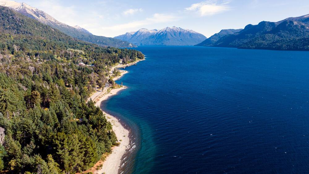 A coastal landscape with a clear blue sea, a sandy beach, dense forests, and snow-capped mountains in the background.