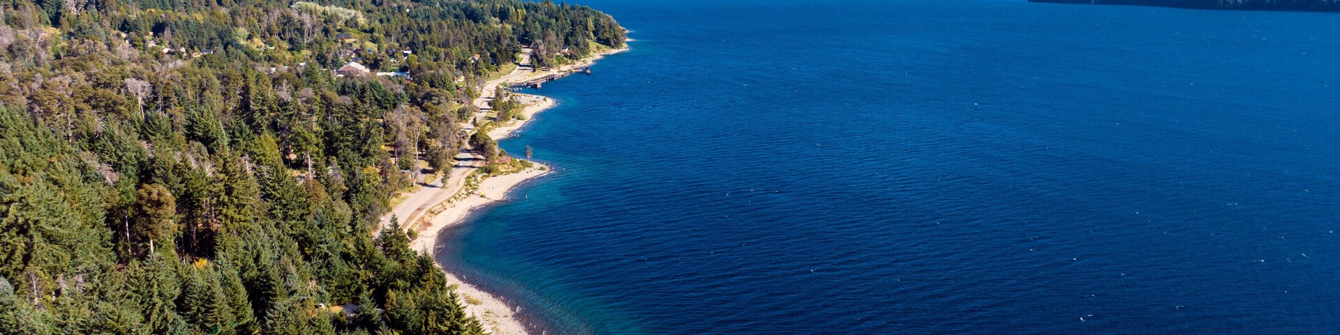 Un paisaje costero con mar azul cristalino, una playa arenosa, bosques densos y montañas nevadas al fondo.