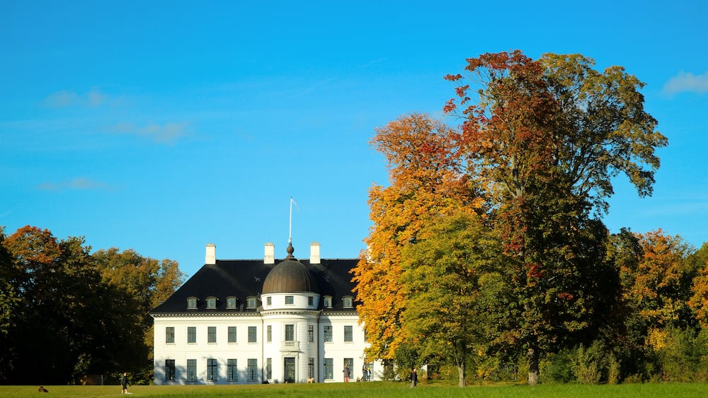 A large white building with a black roof and a dome, surrounded by a grassy field and trees with autumn foliage.