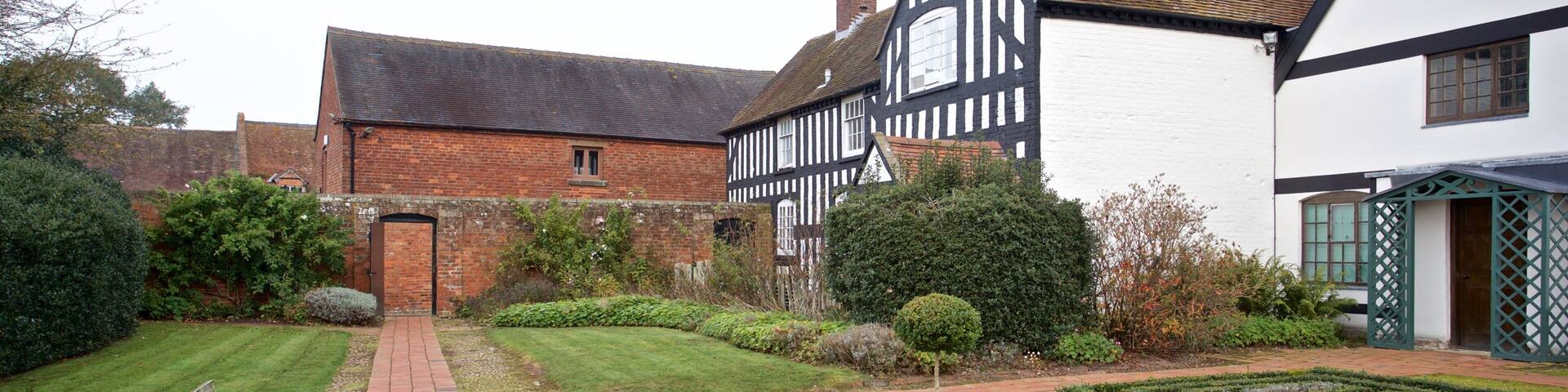 A garden with a brick pathway, a wooden bench, neatly trimmed hedges, and Tudor-style houses.