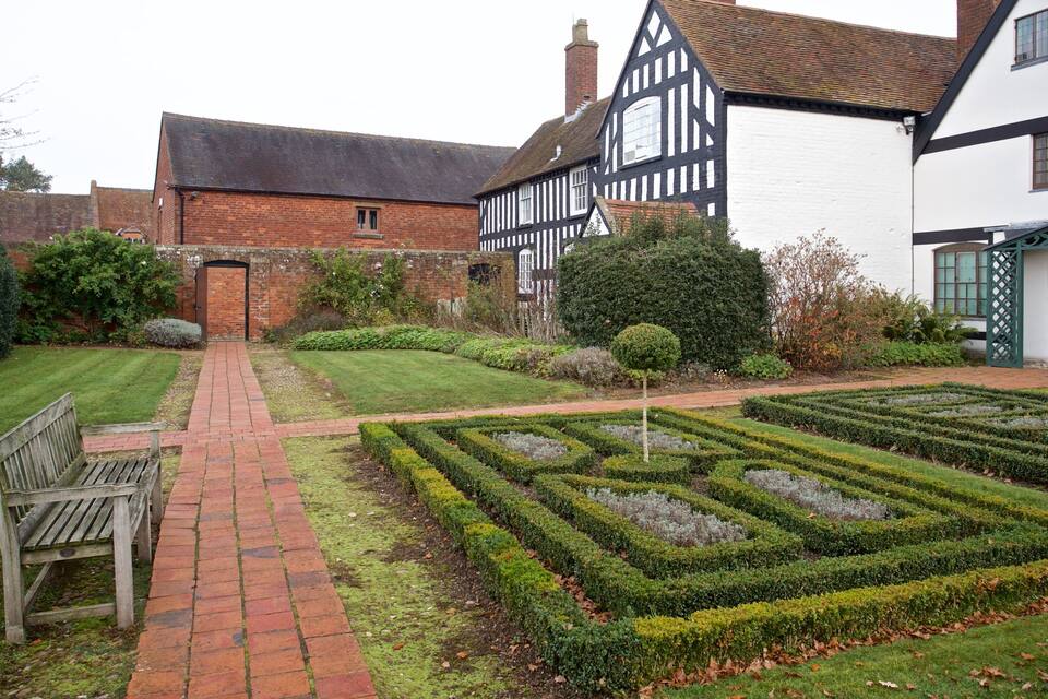 Un giardino con vialetto in mattoni, una panchina in legno, siepi curate e case in stile Tudor.