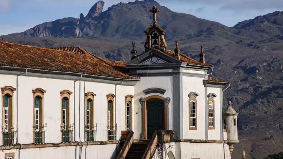 A white building with green windows and a bell tower, set against a mountainous backdrop.