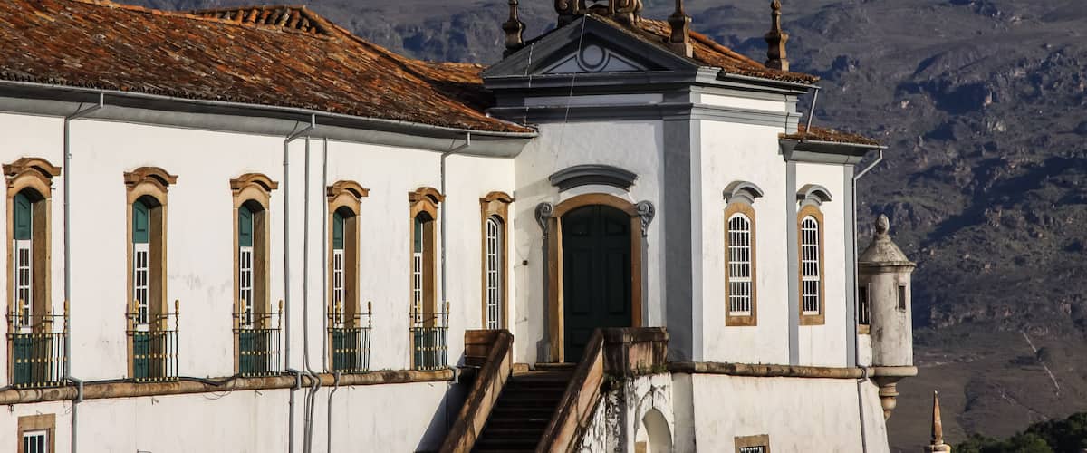 A white building with green windows and a bell tower, set against a mountainous backdrop.