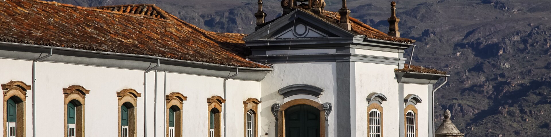 A white building with green windows and a bell tower, set against a mountainous backdrop.
