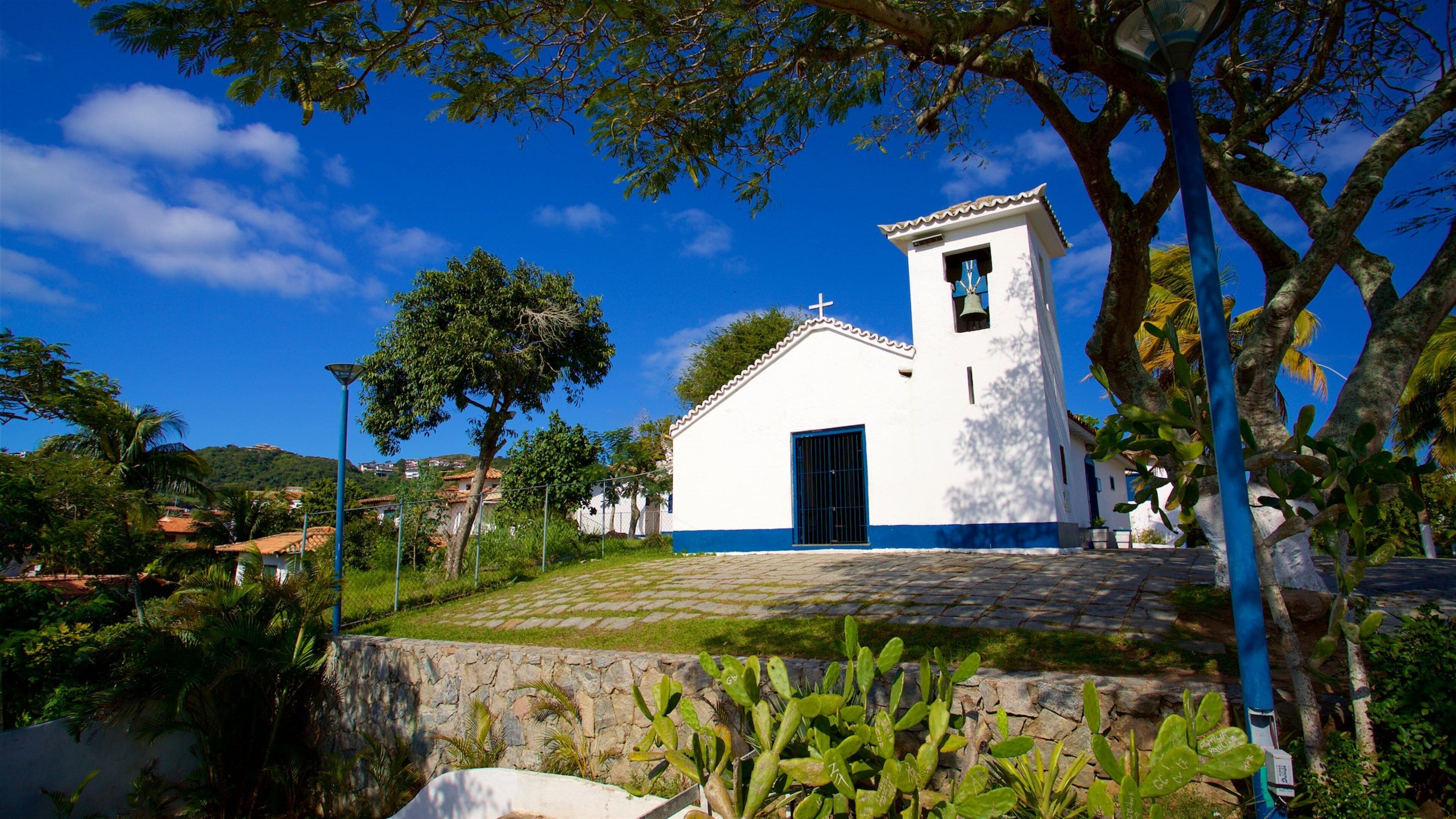 Una iglesia blanca con puerta azul y un campanario, rodeada de vegetación y un cielo azul despejado.