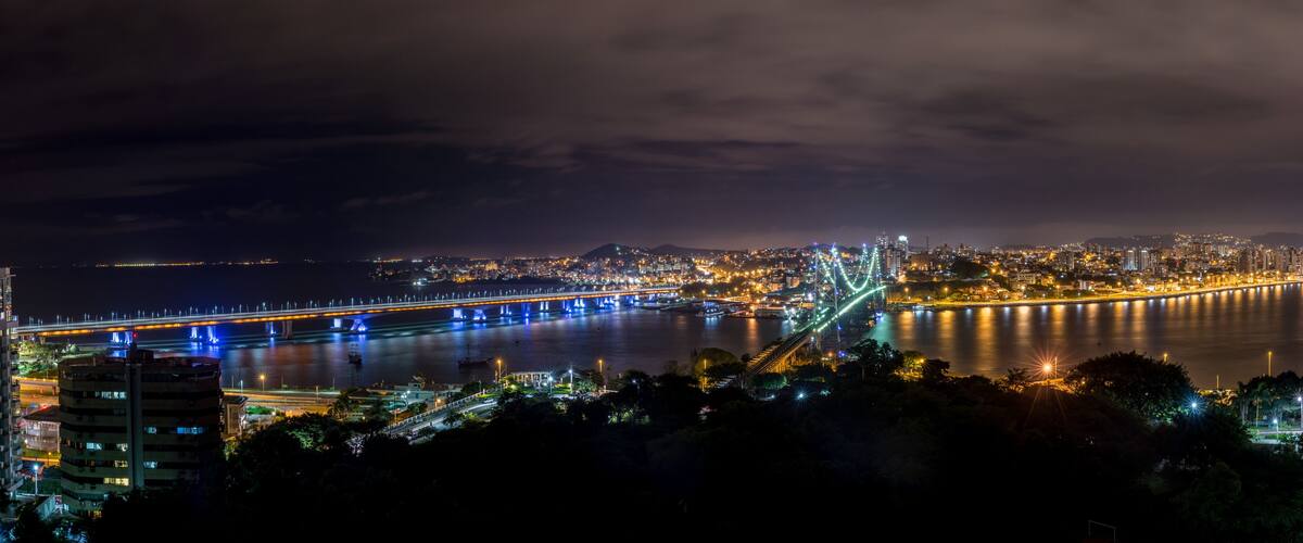 A nighttime cityscape with a brightly lit bridge spanning a river, surrounded by urban buildings and a distant mountain range.
