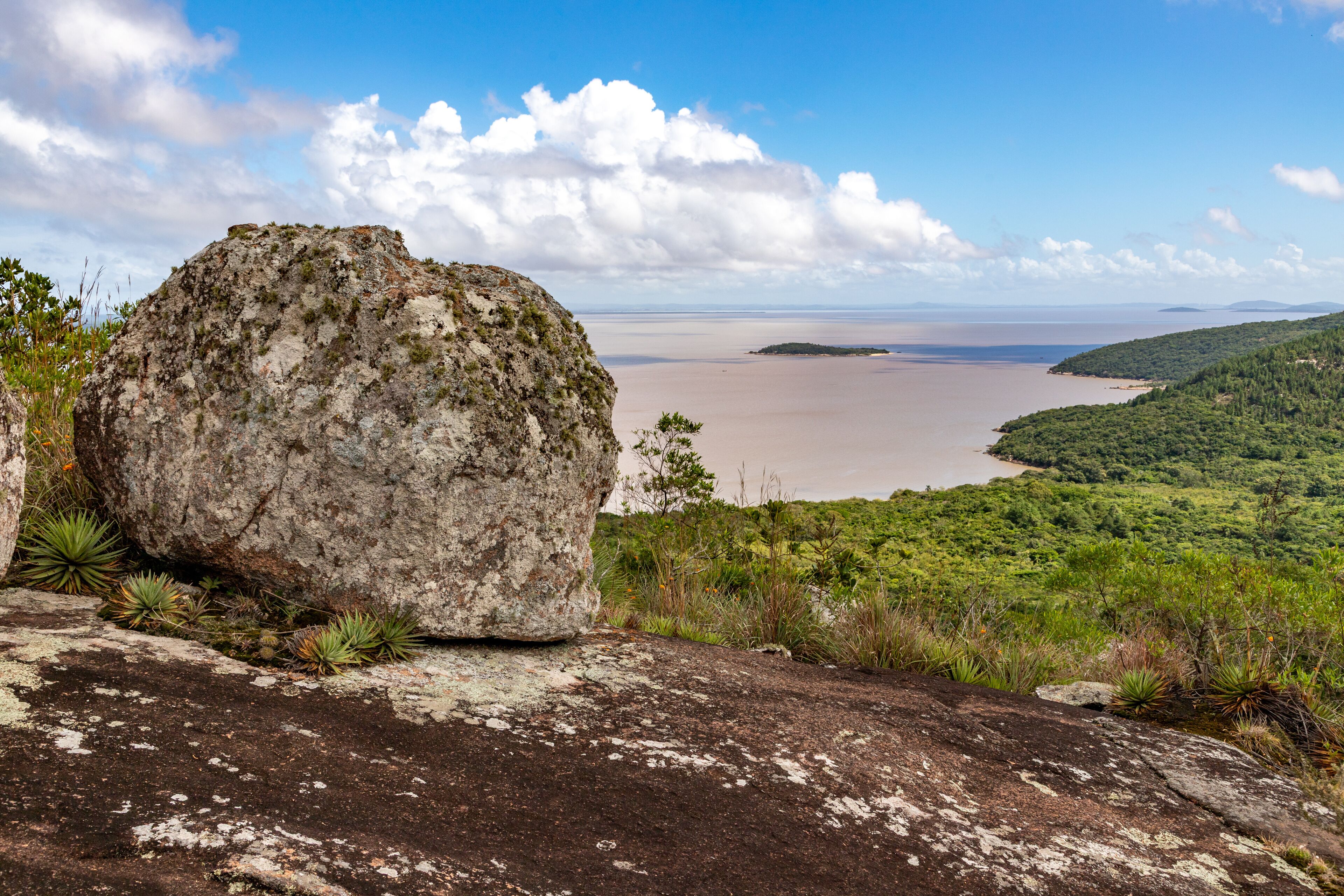 A large rock formation overlooking a calm body of water with a distant island and lush greenery.