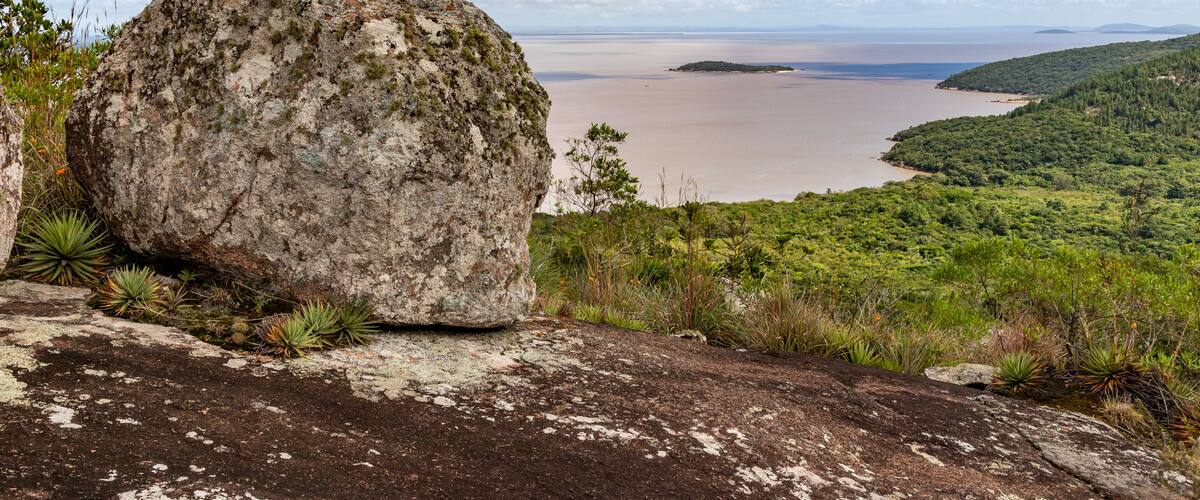 A large rock formation overlooking a calm body of water with a distant island and lush greenery.