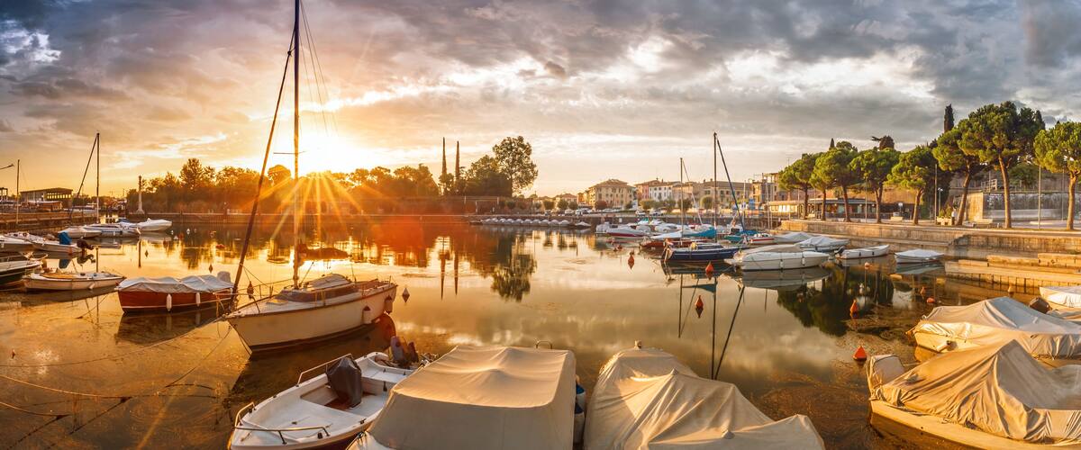 A marina with boats docked, a sunset in the background, and a row of trees along the waterfront.