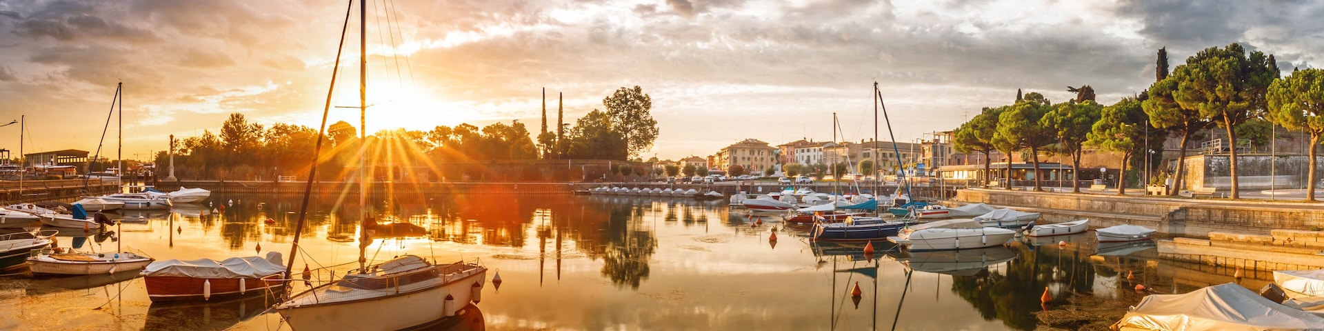 A marina with boats docked, a sunset in the background, and a row of trees along the waterfront.