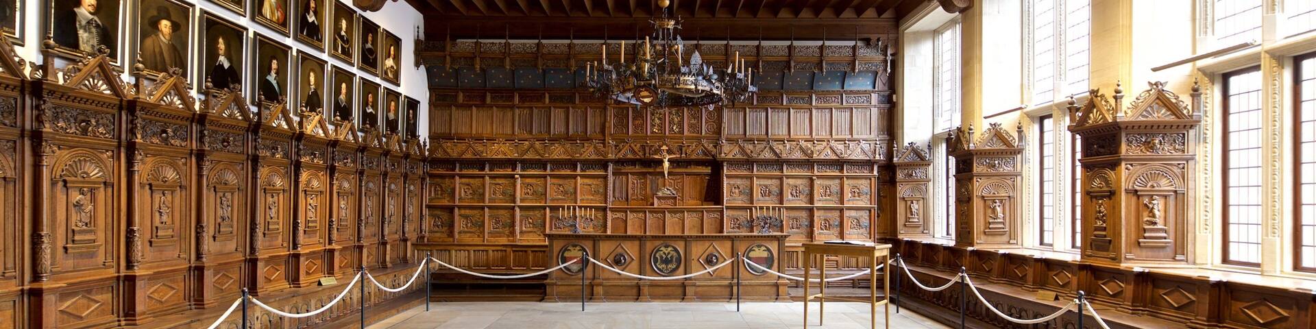 A grand hall with wooden paneling, a chandelier, and framed portraits on the walls.