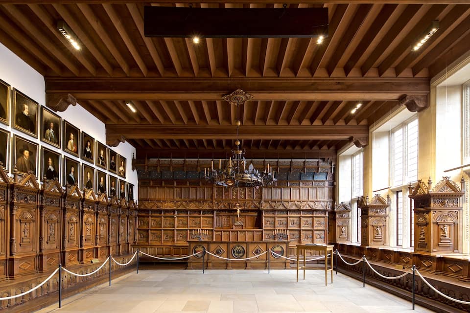 Ein prachtvoller Saal mit Holzwandverkleidung, einem Kronleuchter und gerahmten Porträts an den Wänden.