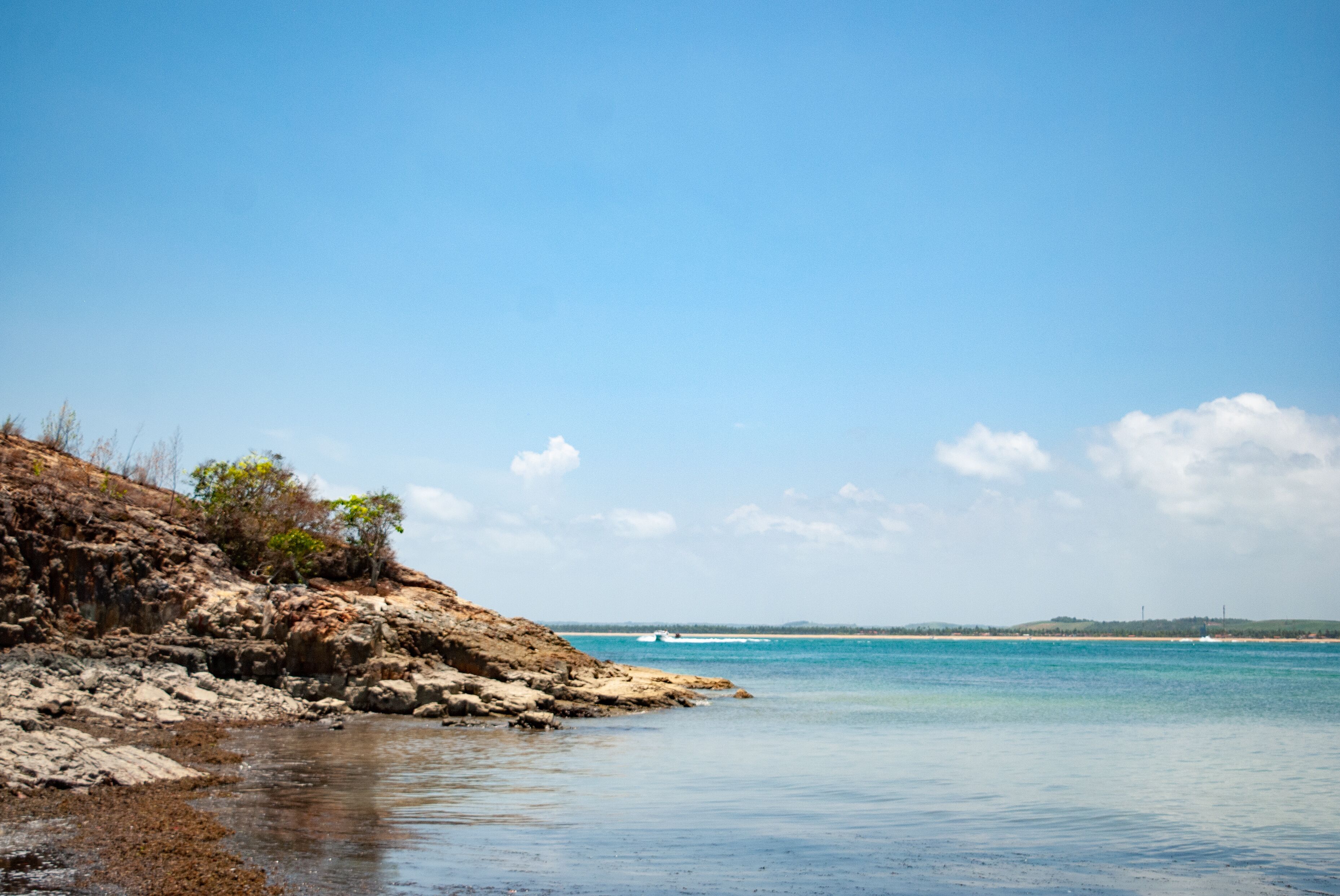 A rocky shoreline with clear blue water and a distant view of a beach.