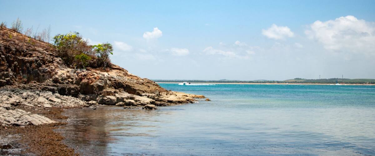 A rocky shoreline with clear blue water and a distant view of a beach.