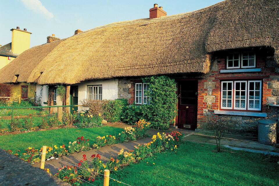A thatched-roof cottage with a brick extension, surrounded by a well-maintained garden with colorful flowers and a wooden fence.