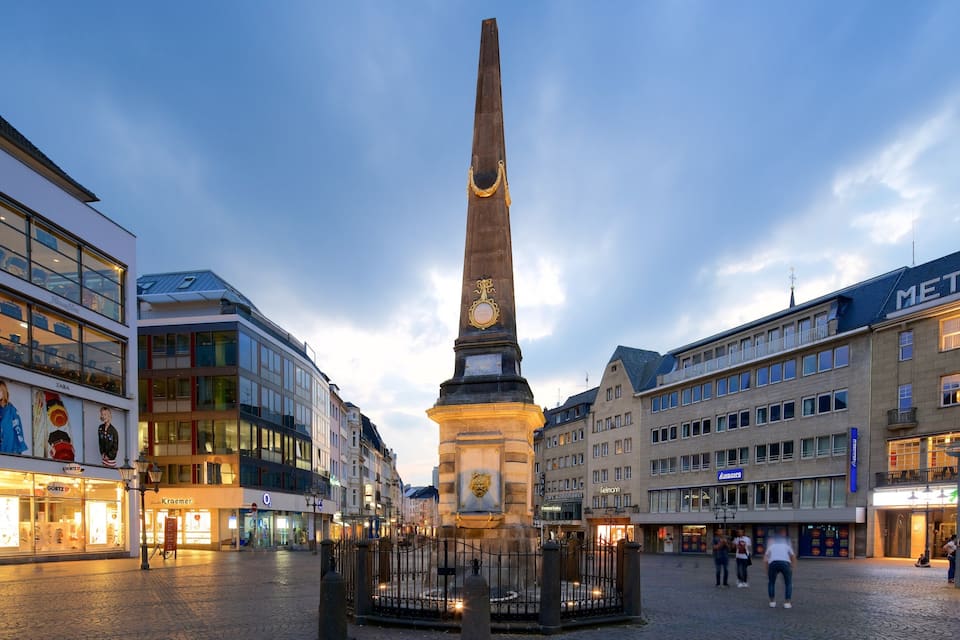 A square with a tall monument in the center, surrounded by modern buildings with illuminated storefronts.