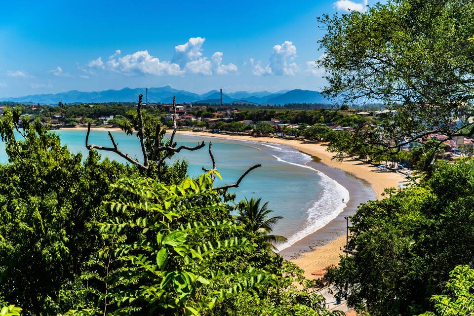 A coastal landscape with a sandy beach, dense greenery, and distant mountains under a clear sky.