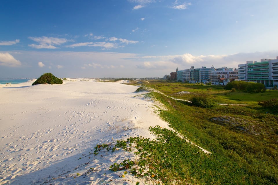 Uma praia arenosa com pegadas, vegetação verde e um complexo de edifícios ao fundo.