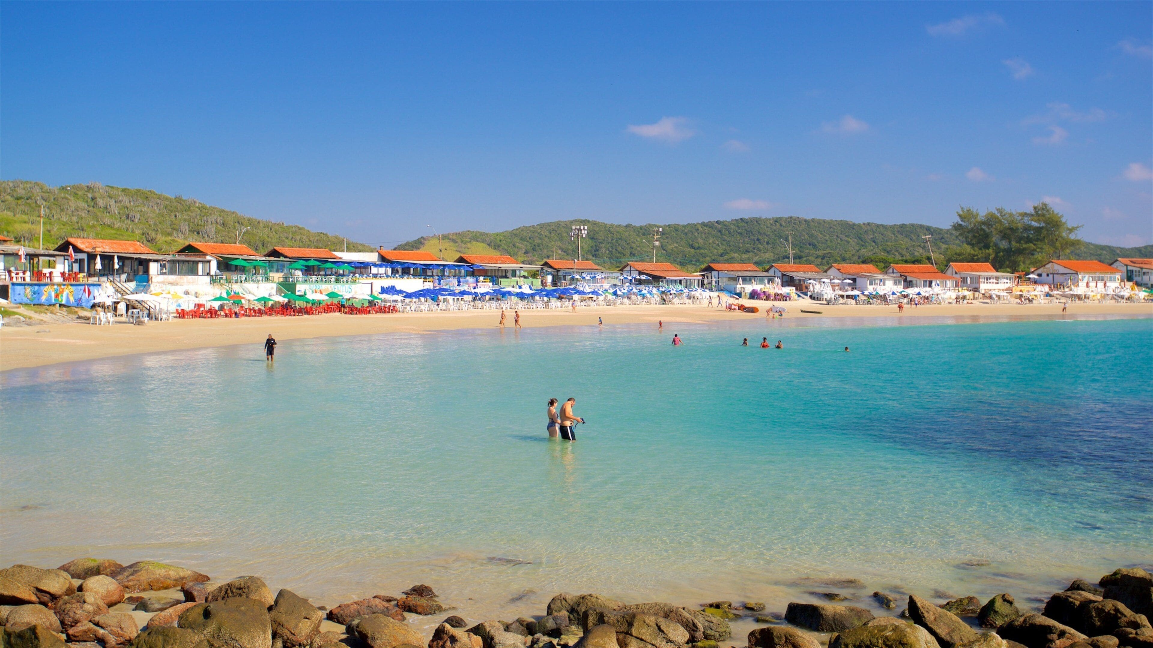 Una playa con aguas turquesas cristalinas, costa rocosa y edificios a la orilla con tejados rojos.