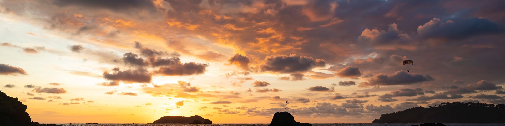 A sunset over a coastal landscape with waves, a rock formation, and a flag on top.
