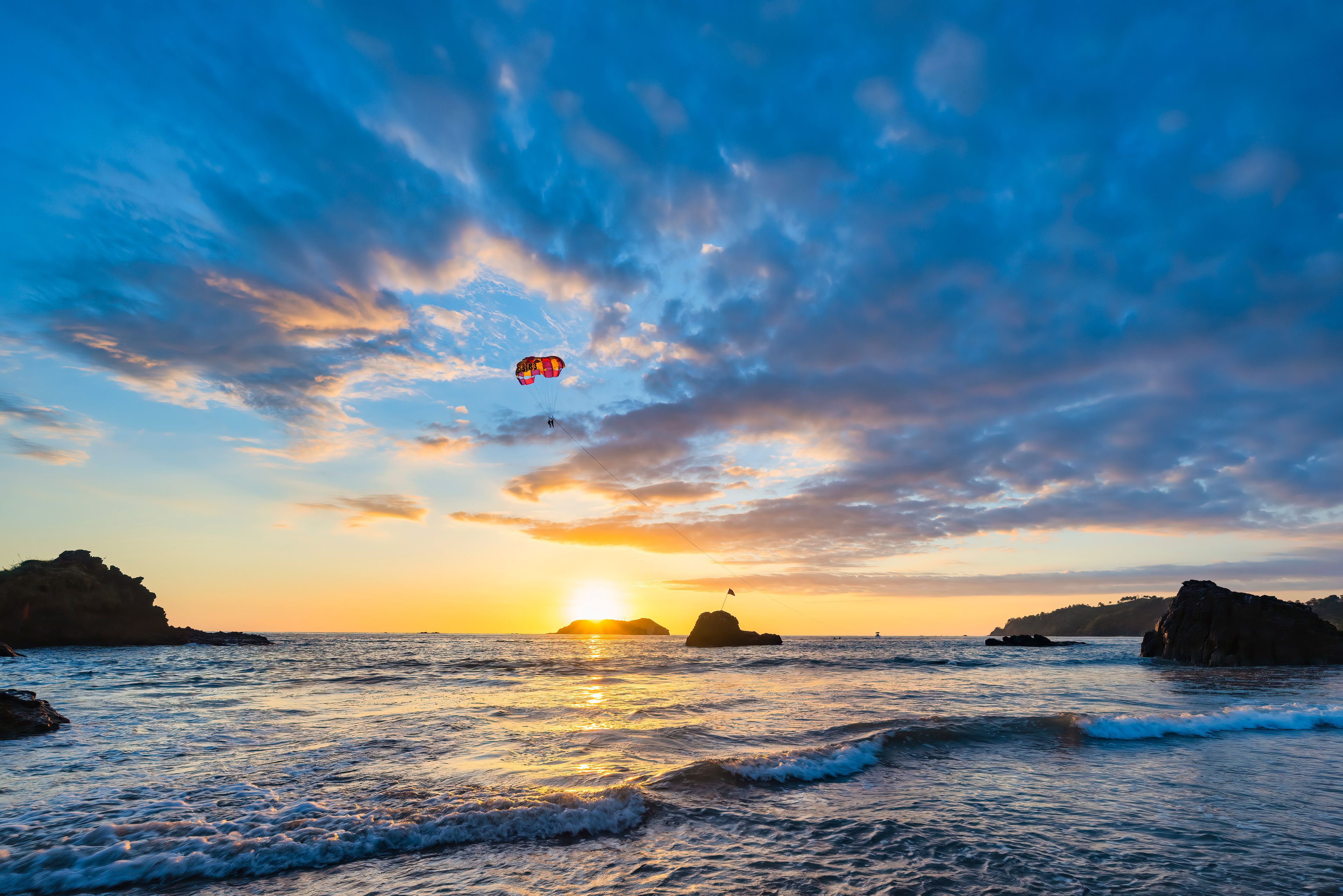 Atardecer sobre el océano con un paracaídas y una bandera en una roca.
