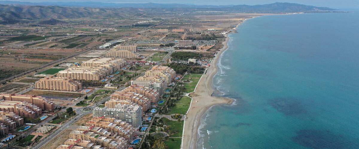 An aerial view of a coastal city with a beach, residential buildings, and distant mountains.