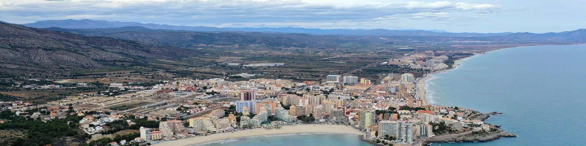 Vista aérea de una ciudad costera con playa, edificios y montañas al fondo.