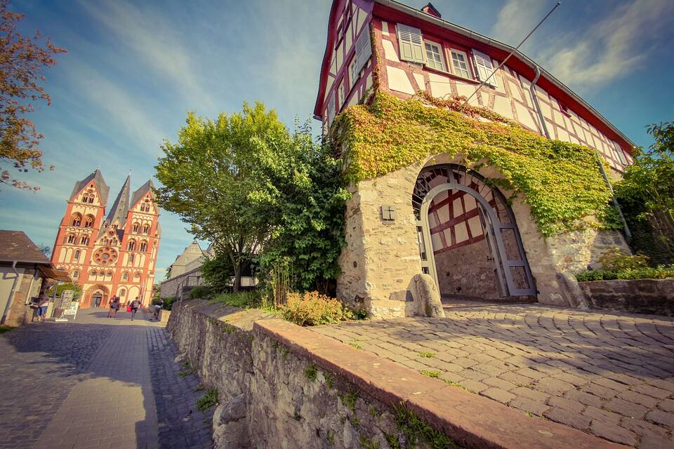 Un bâtiment historique à la façade rouge et blanche, orné d’arches recouvertes de lierre et donnant sur une rue pavée.