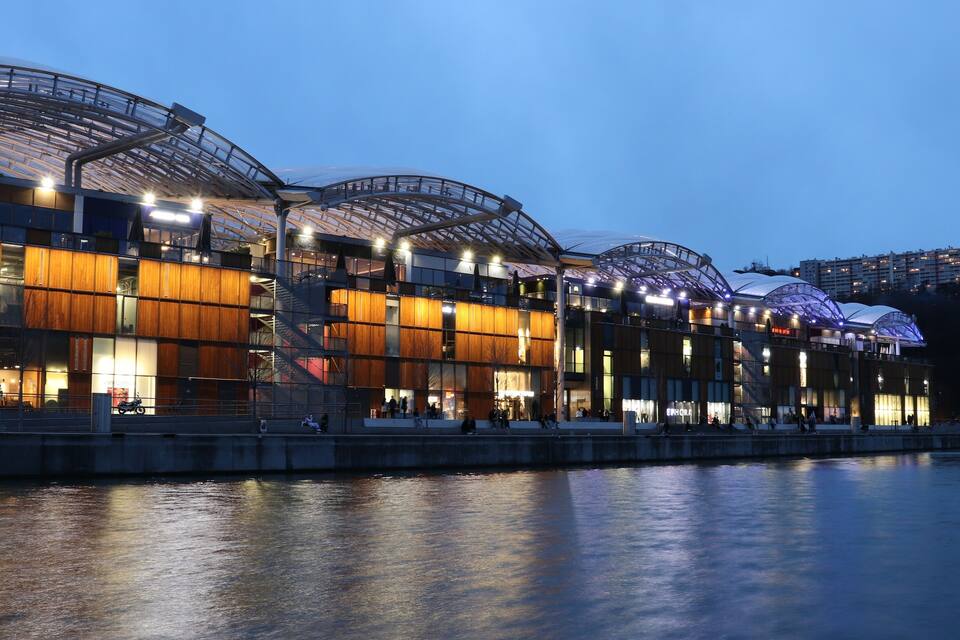 A modern waterfront building with a glass facade and wooden paneling, reflecting on the water.