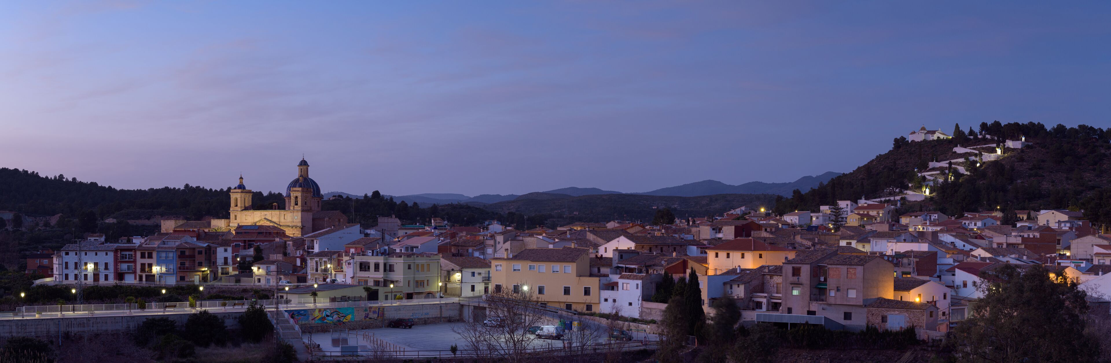 A town at dusk with a prominent church, colorful buildings, and a hill with houses.