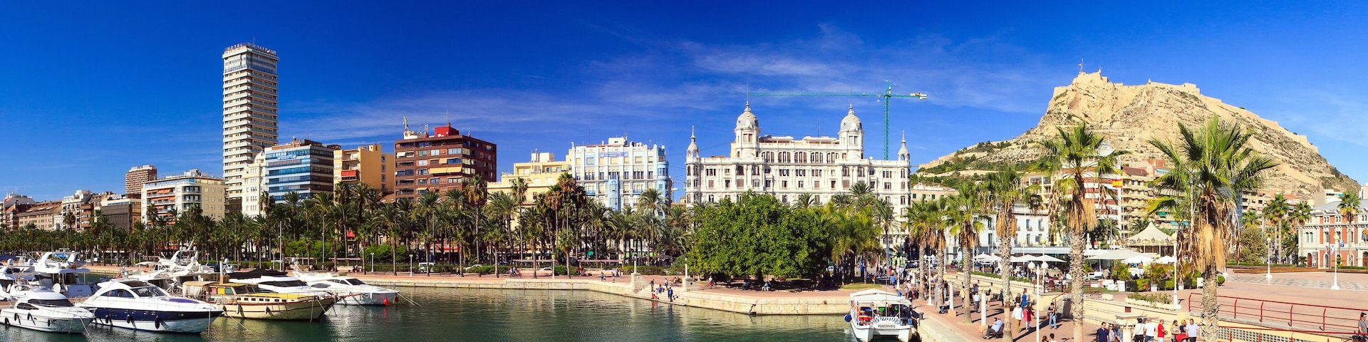 Paisagem costeira com uma colina imponente, edifícios históricos e uma marina com barcos.