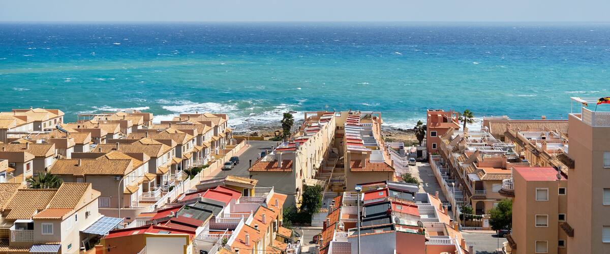 A coastal town with terracotta-roofed buildings, a clear blue sea, and a bird flying in the sky.