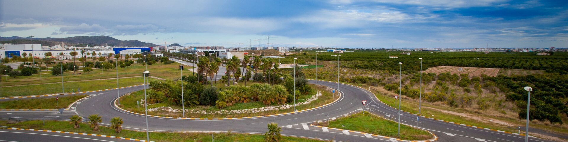 A roundabout with multiple exits, surrounded by greenery and palm trees, with buildings in the background.