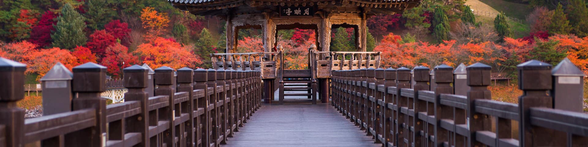 A traditional pavilion on a wooden bridge with autumn foliage in the background.