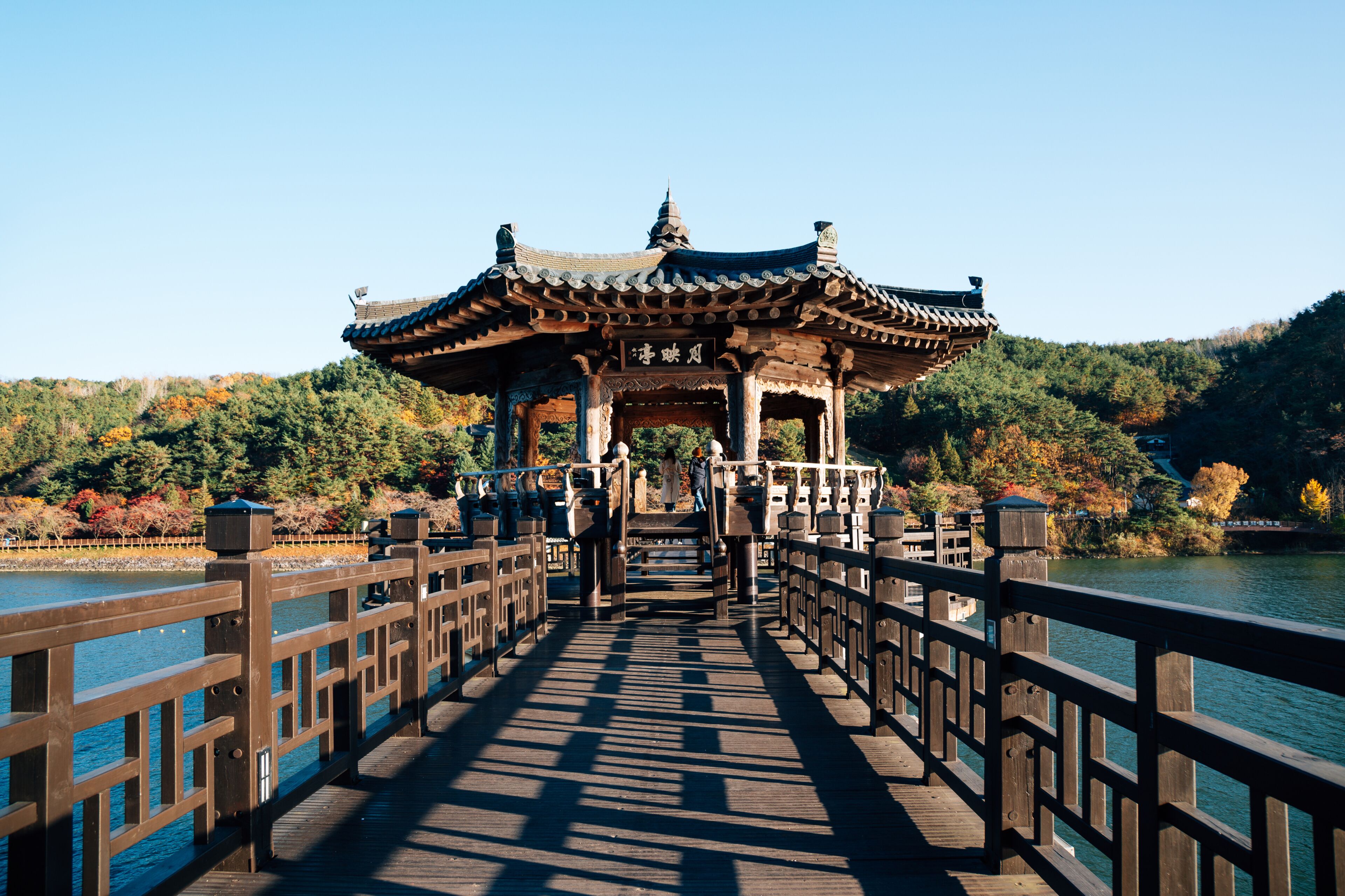 A traditional pavilion on a wooden bridge over a body of water, surrounded by autumn foliage.