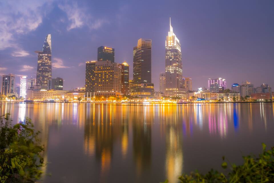 A city skyline at dusk with illuminated buildings reflecting on a calm body of water.