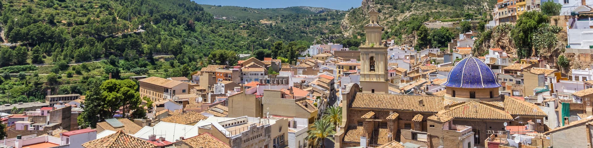 A hillside town with terracotta-roofed buildings, a prominent bell tower, and lush green hills in the background.