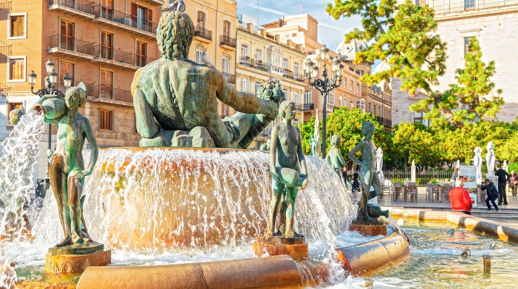 Una fuente de bronce con agua brotando de su base, rodeada de edificios y árboles.